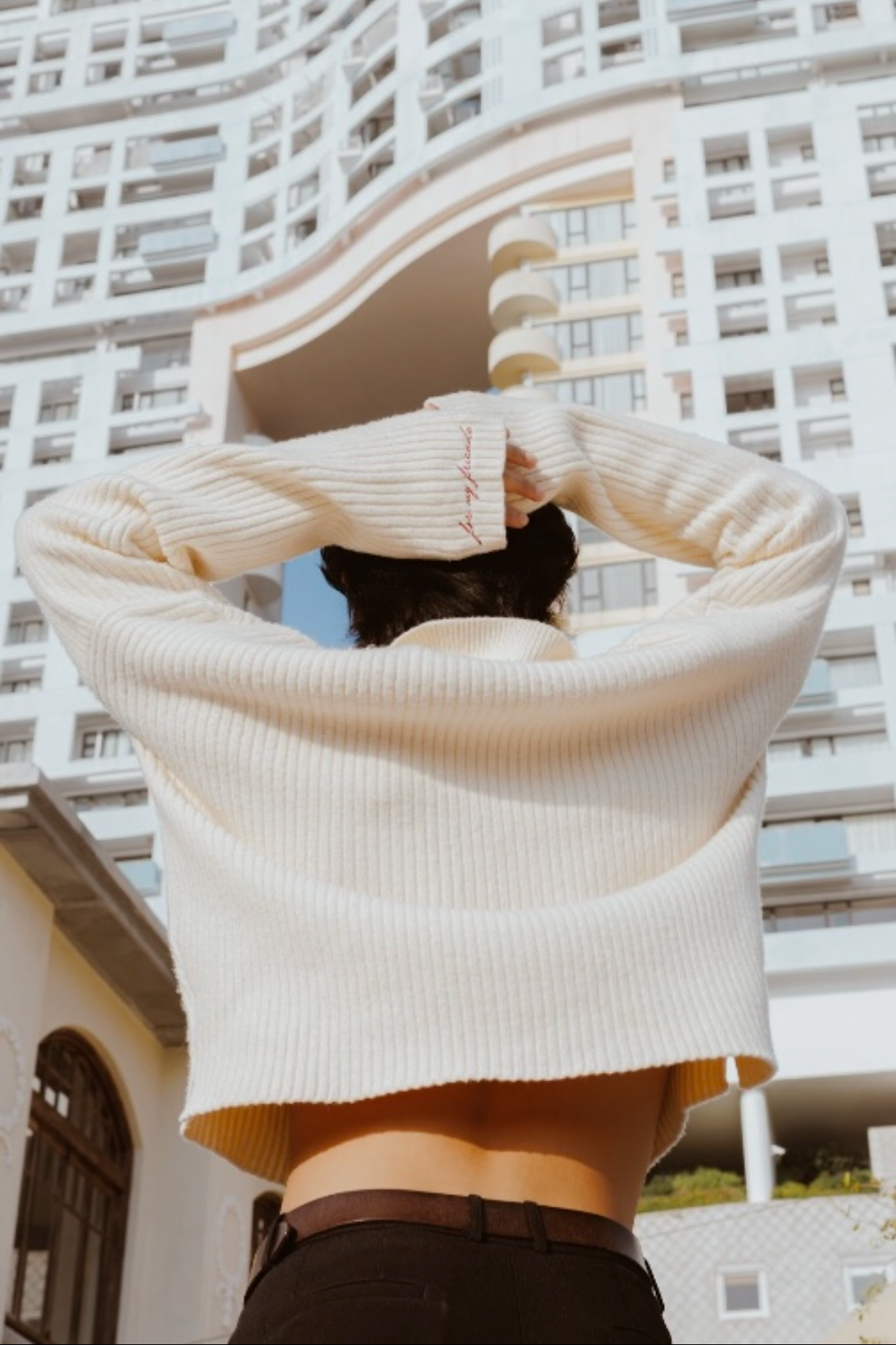 Person wearing a beige sweater with a unique design, standing in front of a modern architectural building.